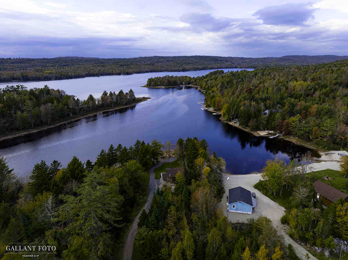 An aerial view captures the serene landscape of Falls Lake Resort, with lush trees dotting the shoreline and gentle hills in the background. The lake's calm waters reflect the surrounding autumn foliage, while the sandy beach and cottages are visible along the water's edge.