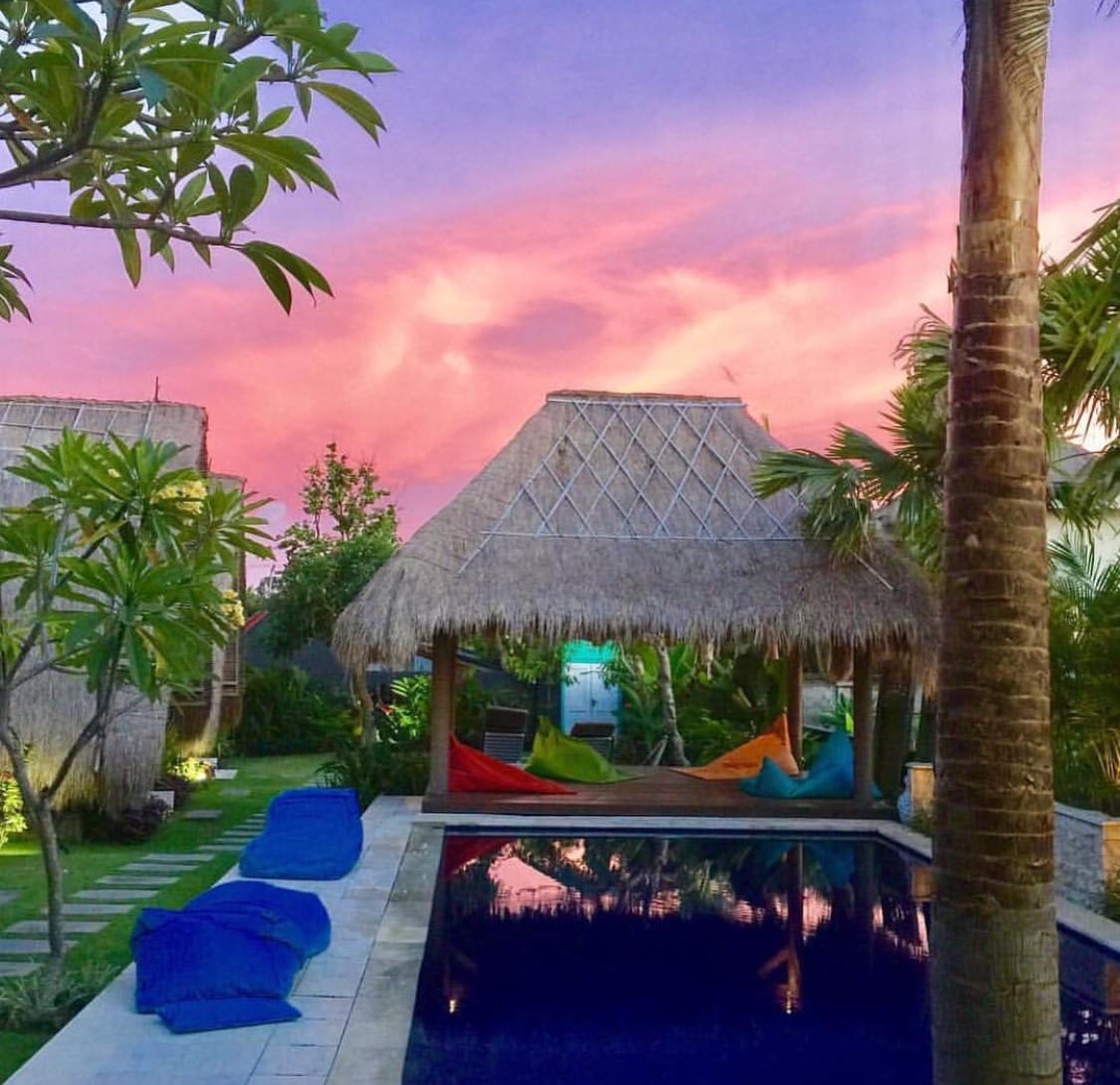 A tranquil outdoor space features a swimming pool reflecting the vibrant colors of a sunset sky. Relaxation is offered by thatched-roof pavilions and colorful hammocks nestled among lush greenery. The pool area is bordered by neatly arranged stone tiles and landscaped garden elements.