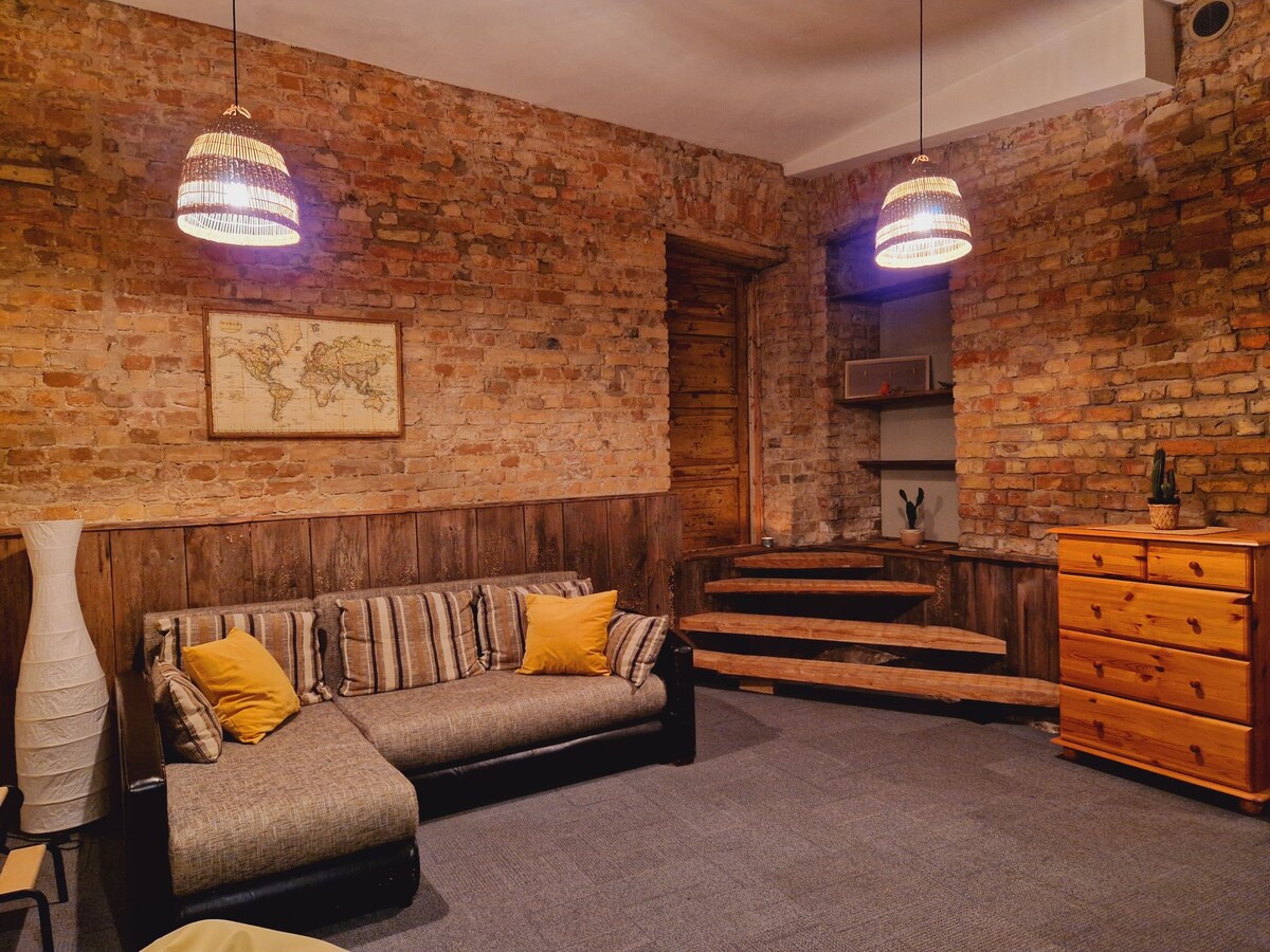 A cozy seating area features a sectional sofa adorned with yellow cushions. Exposed brick walls provide a warm backdrop, while two woven pendant lights hang from the ceiling. A wooden dresser and decorative shelf are subtly positioned in the space.