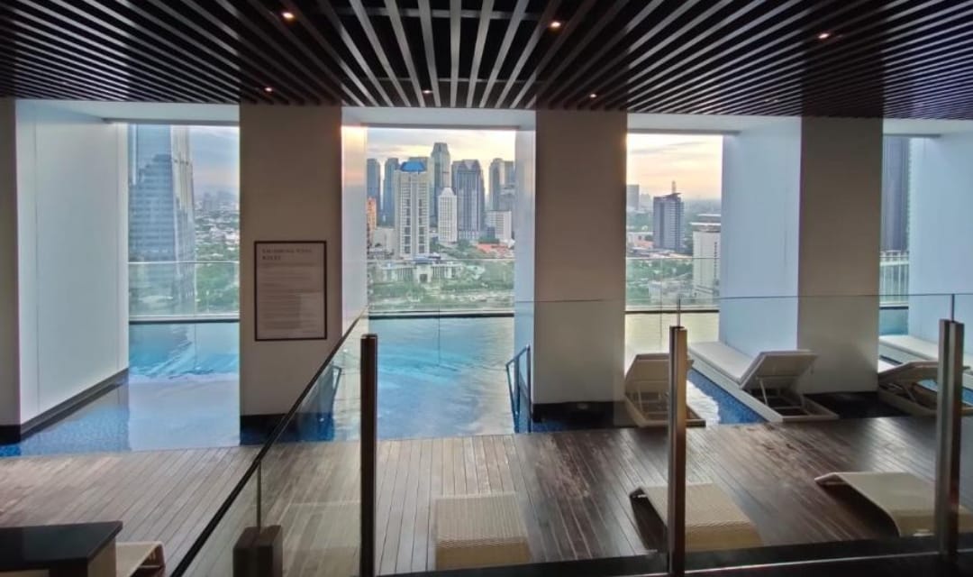 The indoor swimming area features large windows offering views of Jakarta's skyline. Sun loungers are arranged along the wooden deck, with the pool's tranquil water visible in the foreground. Soft lighting enhances the modern architectural design, creating a serene leisure space.