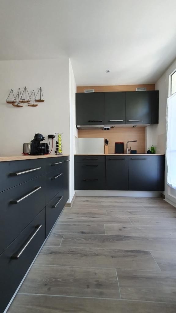 The kitchen features modern dark cabinetry with sleek handles, complemented by a light-colored countertop. Appliances include a coffee maker and kettle, while ample cabinetry provides storage. Natural light floods the space through a window, enhancing the clean and organized layout.