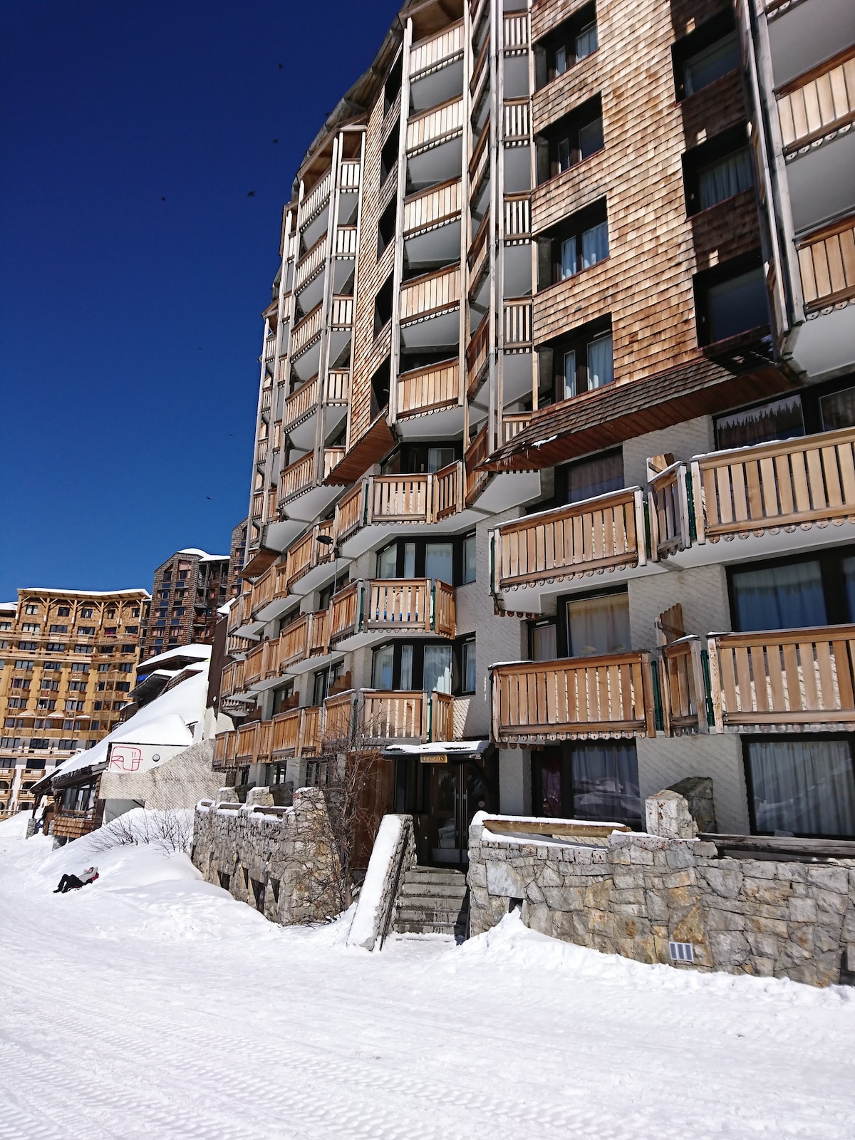 The building showcases a blend of wood and stone exteriors, with multiple balconies visible from each level. Snow-covered grounds surround the structure, highlighting its location in a serene, wintry landscape against a bright blue sky.