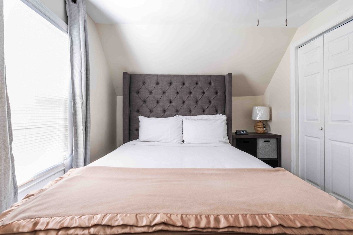 A spacious sleeping area features a tufted gray headboard atop a neatly made bed with white linens. A small nightstand with a lamp stands beside it. Natural light enters through two windows dressed with light gray curtains, and a closet with double doors is visible in the background.