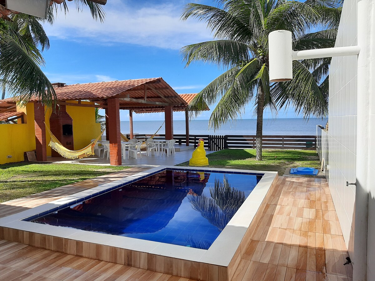A refreshing swimming pool reflects the clear sky, surrounded by manicured grass and two palm trees. A detached quiosque offers shaded seating, with a hammock visible for relaxation. The area presents a seamless view of the ocean beyond.