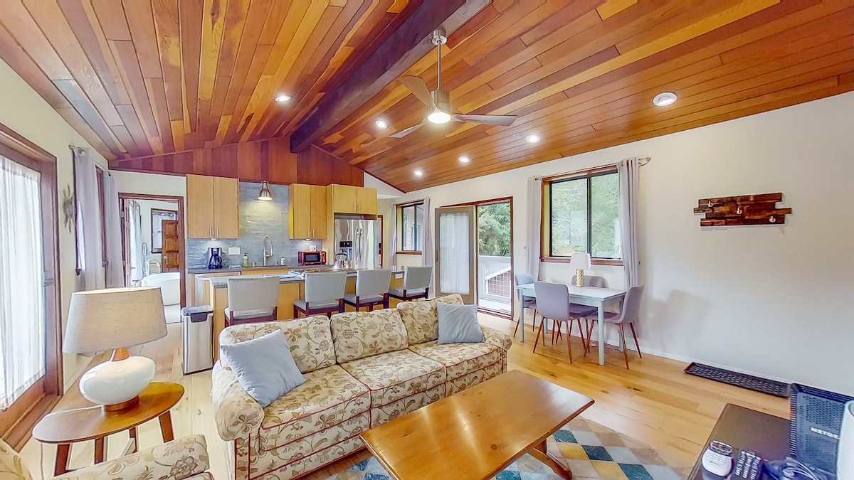 An open living space features a comfortable sofa with decorative cushions, a wooden coffee table, and a dining area with chairs. Large windows let in natural light, while a sleek kitchen is visible in the background, showcasing modern appliances and finishes.