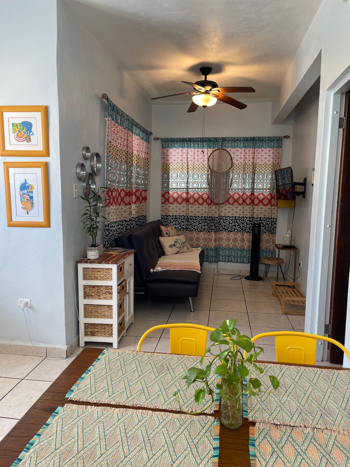 A bright living-dining area features a full-size sofa bed and colorful patterned curtains. A small table with chairs sits in the foreground, while light wood accents are visible throughout. A ceiling fan and TV can be seen, enhancing the room's functionality.