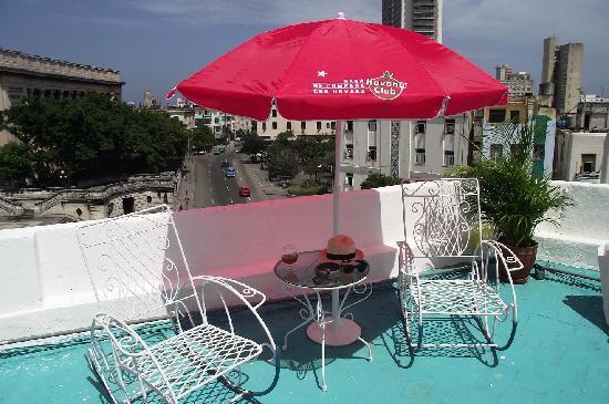 A rooftop terrace is presented, featuring a red umbrella providing shade over a small round table. Two white metal rocking chairs surround the table. The vibrant turquoise floor contrasts with the greenery of nearby plants and the urban backdrop of the city streets below.