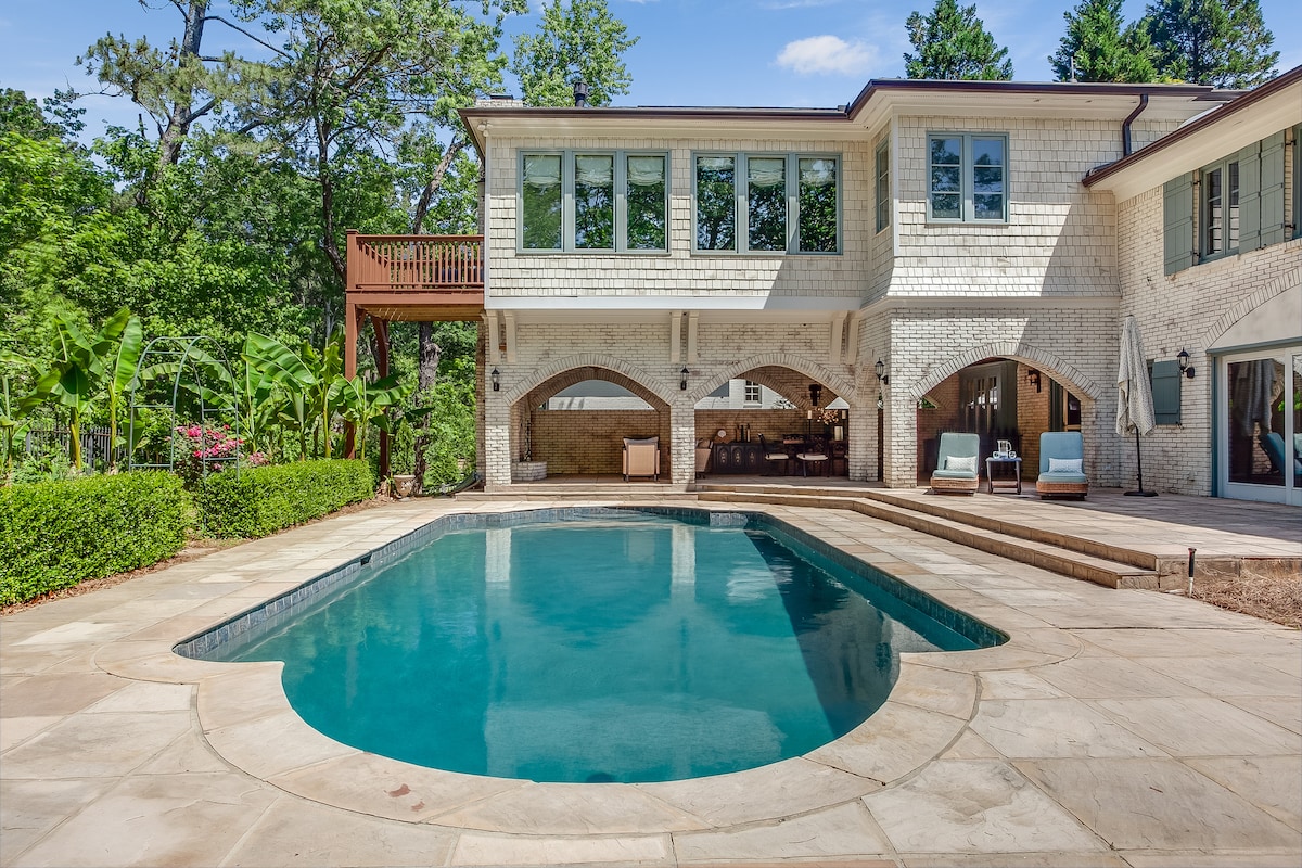 A serene outdoor space features a gentle pool surrounded by a stone patio. The walls of the house, adorned with large windows, reflect natural light and greenery. An upper balcony is visible, enhancing the private atmosphere of the backyard.