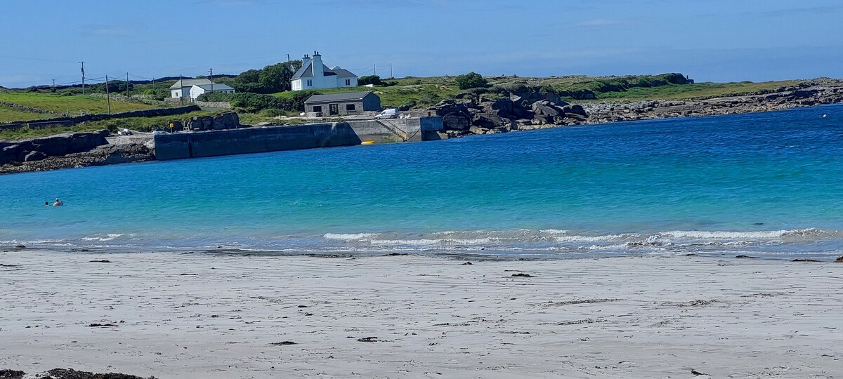 A serene beach scene features a soft sand shore leading to clear blue waters. In the distance, a white building is situated on a grassy hill, with rugged coastline visible to the side. Gentle waves lap at the shore under a bright blue sky.