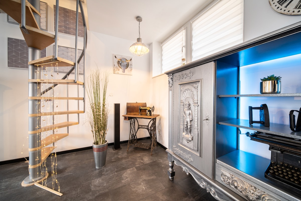 A modern interior features a spiral staircase leading to an upper level. A vintage sewing machine is placed on a wooden table, while a silver cabinet displays decorative items against a backdrop of natural light from windows. Green plants add a touch of nature.