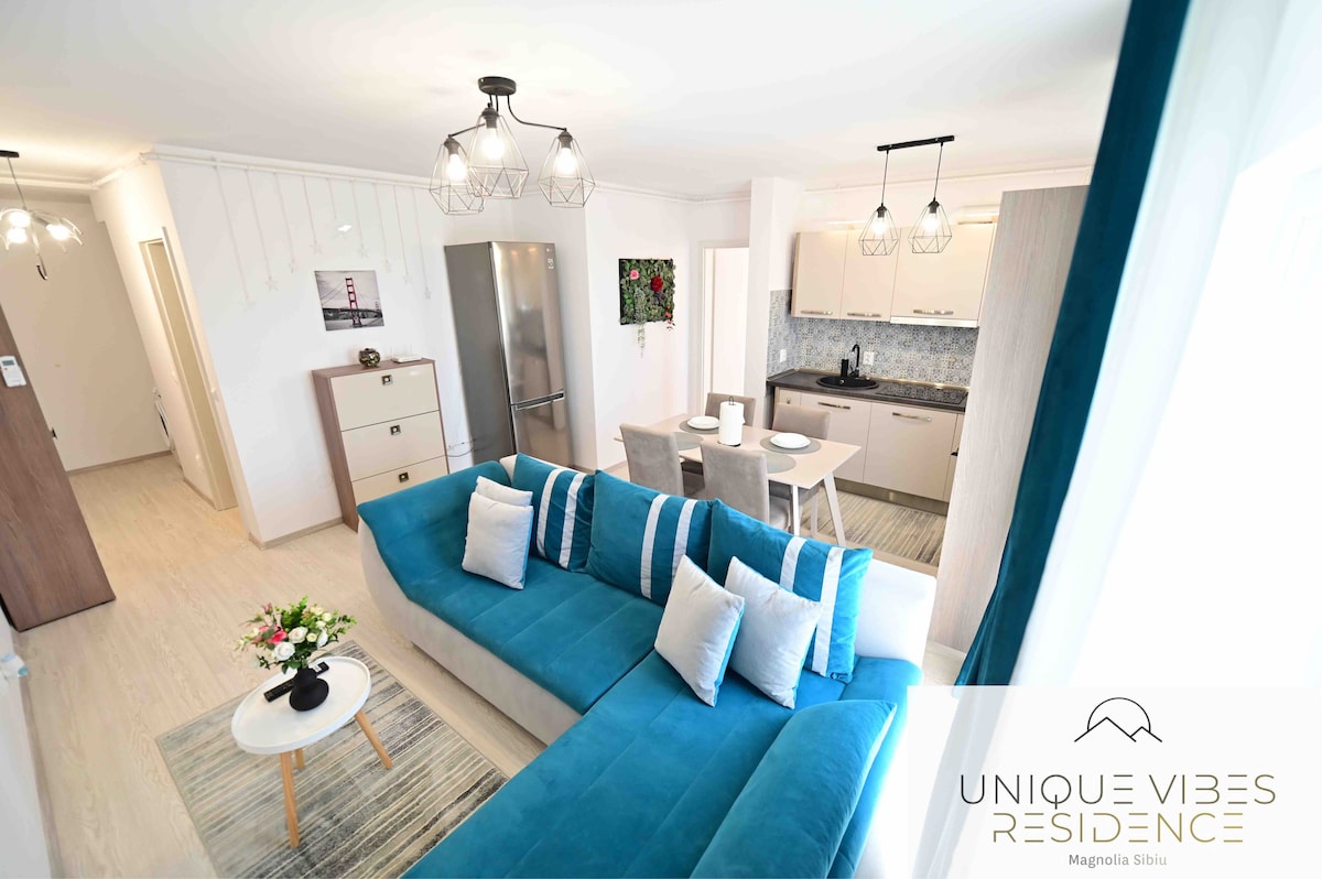 An open-plan living area is presented, featuring a spacious blue sectional sofa and a coffee table. A dining area for four is visible, alongside a modern kitchen with sleek cabinetry and stainless steel appliances. Natural light fills the room, creating a bright, inviting atmosphere.