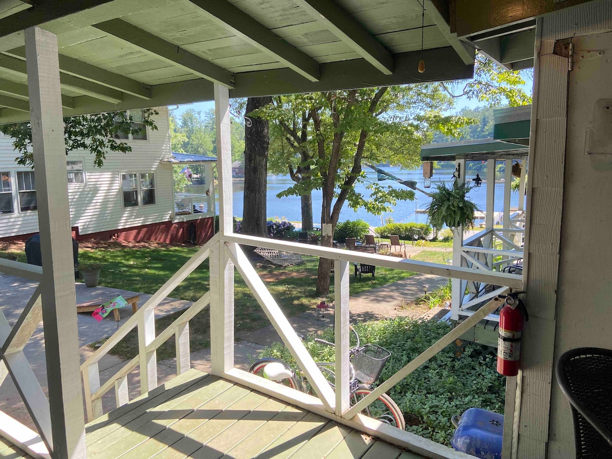 A view from the porch reveals a serene lakeside scene, with green trees framing a tranquil body of water. A grassy area is dotted with picnic tables and lounge chairs, inviting relaxation. Additional outdoor activities are suggested by visible bicycles and canoes nearby.
