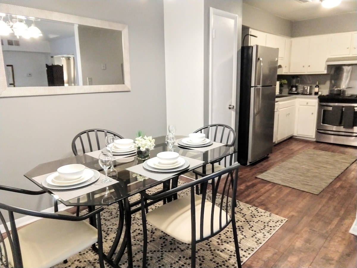 A dining area features a glass table surrounded by four black chairs, set for a meal with plates and glasses. The kitchen is visible in the background, equipped with modern appliances and cabinetry. A neutral-toned area rug complements the warm wood flooring.