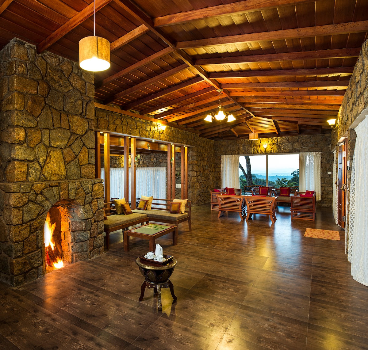 The spacious common lobby features natural stone walls and a wooden ceiling. An indoor fireplace adds a warm touch. Comfortable seating arrangements with colorful cushions invite relaxation. Large windows provide expansive views of the surrounding landscape, enhancing the connection with nature.