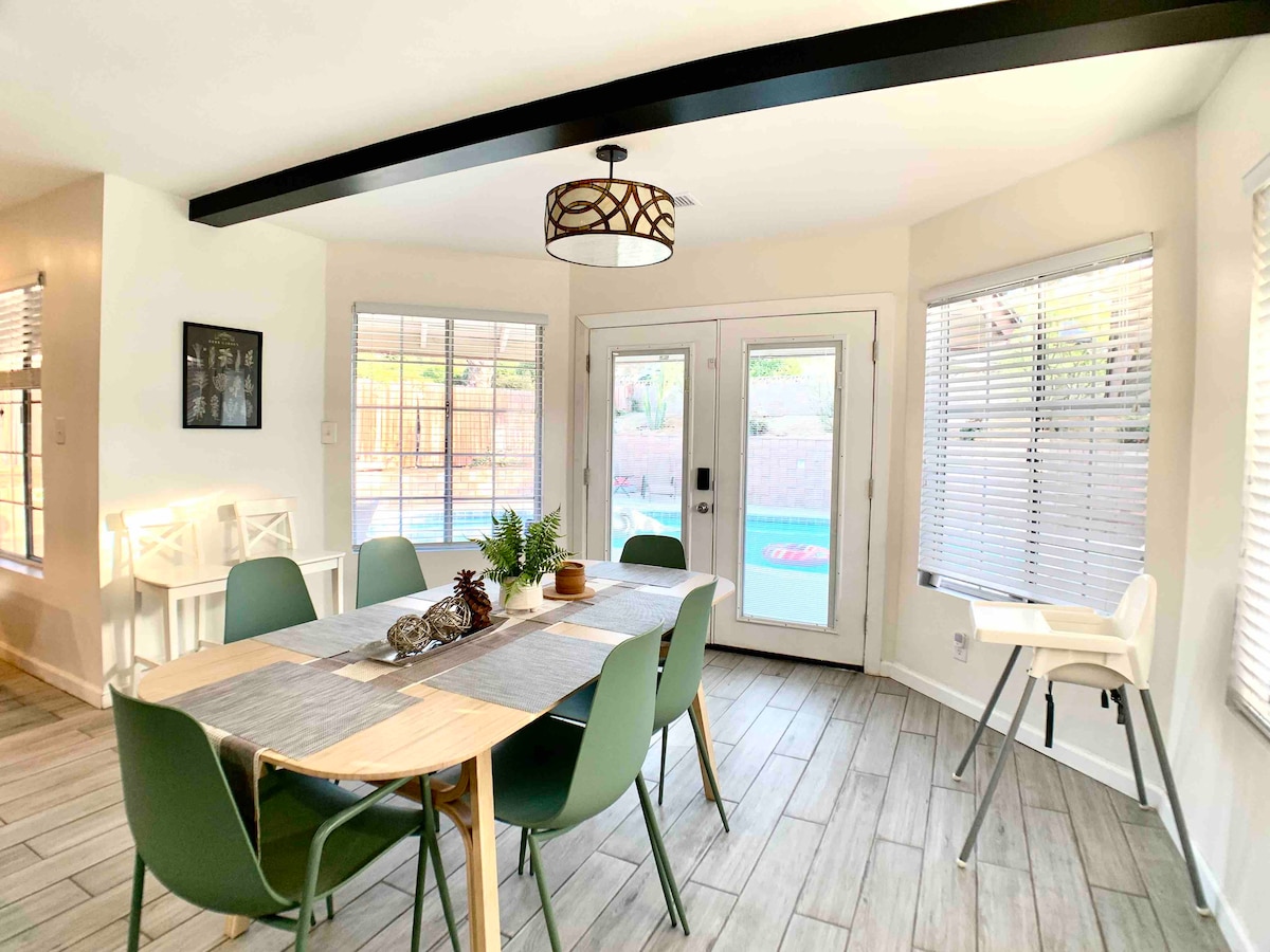 A dining area features a wooden table surrounded by six green chairs. Natural light enters through large windows with blinds. A high chair is positioned at the end of the table. Double doors provide access to the outdoor space, revealing a glimpse of the pool.
