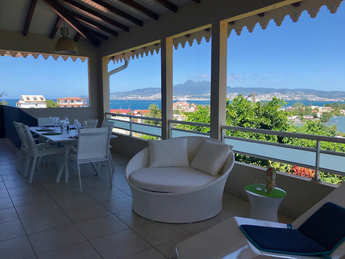 An outdoor terrace features a round white seating area with a small table, surrounded by a spacious dining setup for six. Panoramic views of the bay and distant mountains can be seen through large open windows, illuminated by natural light.