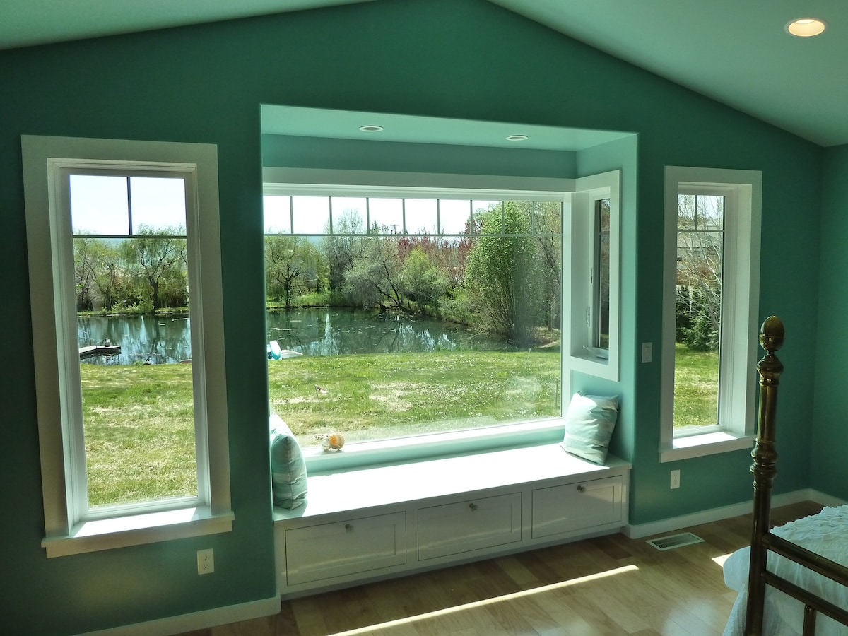 A serene view of the outdoor landscape is framed by three windows in a light blue wall. The scene showcases a peaceful pond surrounded by greenery, with soft natural light illuminating the space. A cushioned window seat provides an inviting spot to relax and enjoy the view.