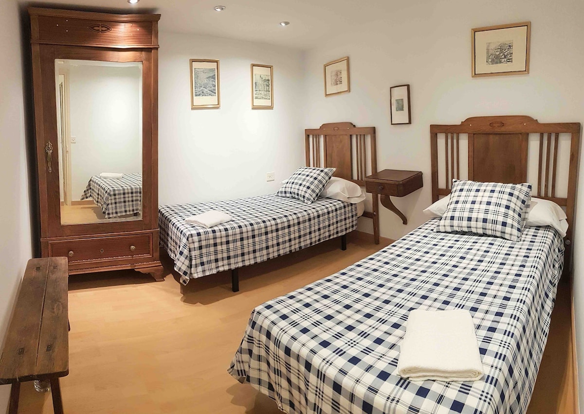 Two single beds with checkered bedding are positioned in a well-lit room. A wooden mirror and a rustic bench provide functionality, while framed artwork adorns the walls, adding a touch of character to the space.