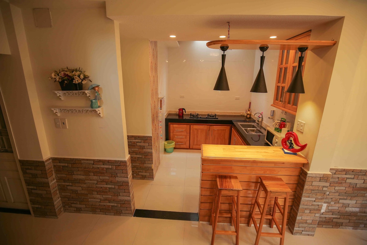 A well-equipped kitchen is showcased with wooden cabinetry and modern appliances. Two wooden stools are positioned at a small bar area. Pendant lights hang above the counter, providing ample illumination. The space is designed with a blend of tile and stone accents for a cozy feel.