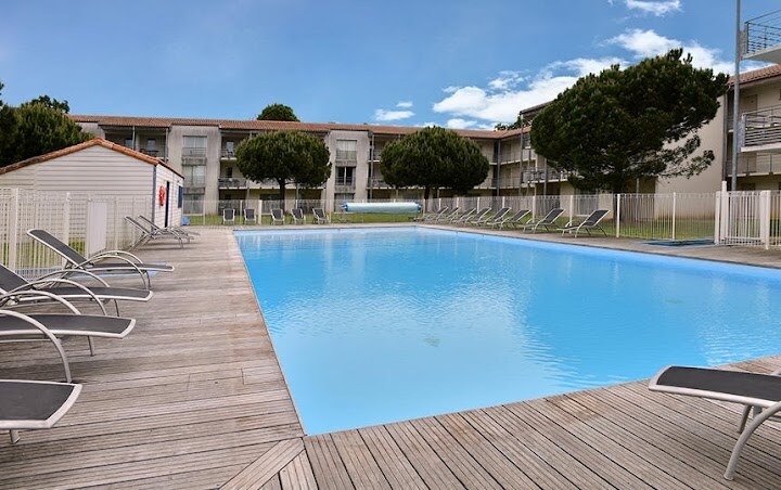 A spacious outdoor pool is surrounded by a wooden deck, featuring several loungers arranged along its perimeter. Lush trees provide shade nearby, and a low fence encloses the pool area. The building's exterior is visible in the background against an open sky.