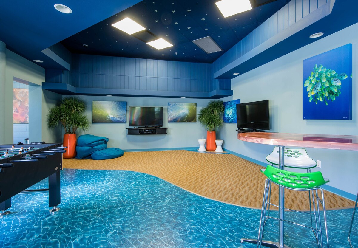 A vibrant game room features a sandy texture flooring with a playful blue accent. Multiple televisions are mounted on the wall, and inflatable seating provides a comfortable area. An artistic piece of sea life decorates the wall, enhancing the room's lively atmosphere.