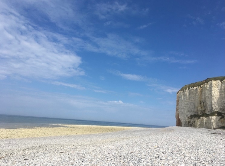 A wide expanse of pebbled beach stretches towards the horizon, meeting calm waters under a clear blue sky. The cliffs rise majestically in the background, showcasing their natural texture and color, while a gentle shoreline invites exploration.