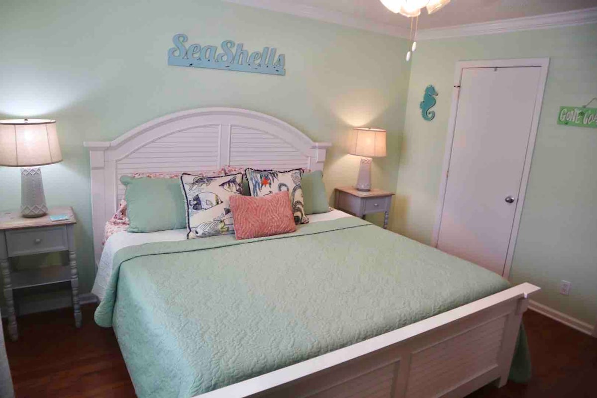 A serene bedroom features a spacious bed adorned with a mint green coverlet and decorative pillows. Soft lighting is provided by two bedside lamps. The light mint walls enhance the beach-themed decor, complemented by wall art that reads 'SeaShells.' A closet door is visible in the background.