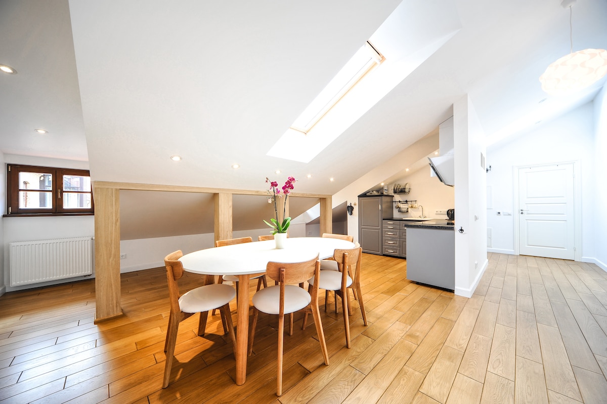 A well-lit dining area features a round table surrounded by six wooden chairs. Natural light streams through skylights and windows, highlighting the warm wooden flooring. In the background, a modern kitchen is visible with sleek cabinetry and cooking appliances.