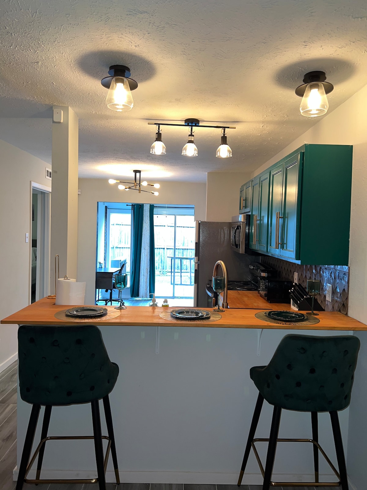 The kitchen features modern green cabinetry and a sleek wooden counter with two high-backed stools. Above, stylish pendant lights illuminate the space. A glass sliding door provides access to the outdoor patio, allowing natural light to stream in.