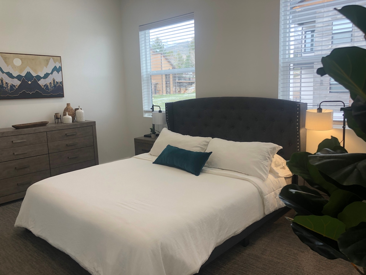A well-appointed bedroom features a spacious bed adorned with crisp white linens and a decorative pillow. Natural light enters through two windows, illuminating the neutral walls and accentuating a stylish artwork piece on the wall. A dresser and bedside lamp complete the room's layout.