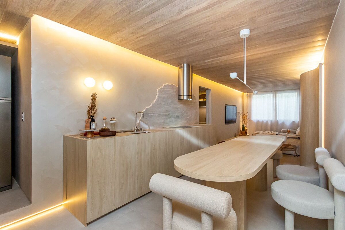 A minimalist kitchen and dining area is presented with light wood cabinetry and a spacious dining table. Soft lighting creates a warm ambiance, complemented by modern appliances and a large window that allows natural light to fill the space.