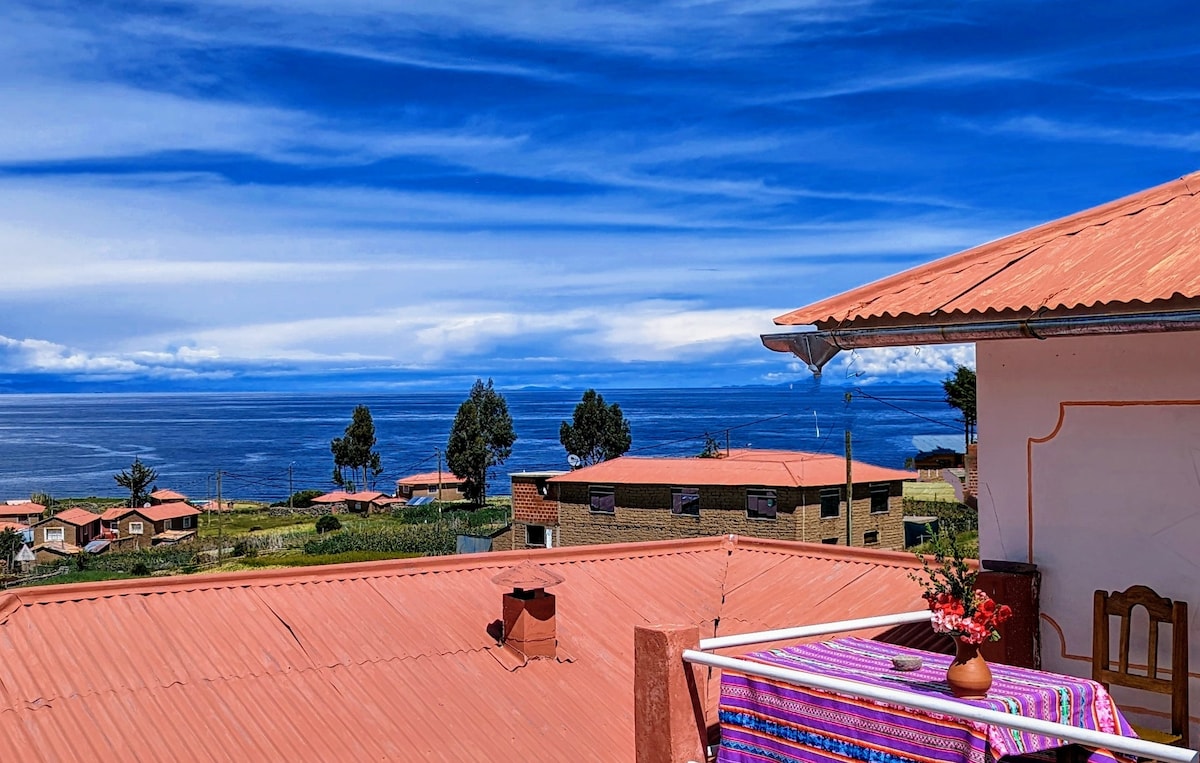 A rooftop terrace features a table adorned with a colorful tablecloth and a small vase of flowers. The expansive view of Lake Titicaca and surrounding houses under a bright blue sky creates an inviting atmosphere for relaxation.