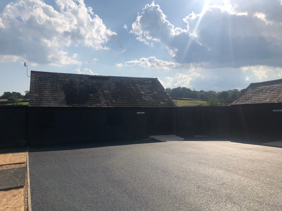 A newly renovated barn exterior is visible, showcasing a dark wood facade under a partly cloudy sky. Sunlight streams from above, illuminating the surrounding landscape, which features green hills in the distance. A smooth, paved area is situated in front of the barn.