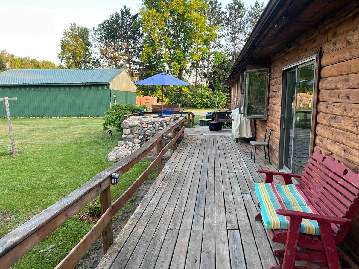 A wooden patio extends from the log home, featuring a red chair with colorful stripes. A stone fire pit is visible in the background, surrounded by seating areas beneath a large blue umbrella. Lush green grass surrounds the patio, set against a backdrop of trees.
