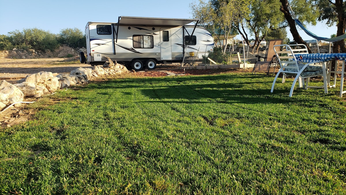 A travel trailer is parked on a well-maintained grassy area, with a shaded awning providing shelter. Nearby, white outdoor chairs are positioned around a blue-checkered table, creating a cozy spot for relaxation. Surrounding landscaping features natural stone borders and trees, enhancing the peaceful setting.