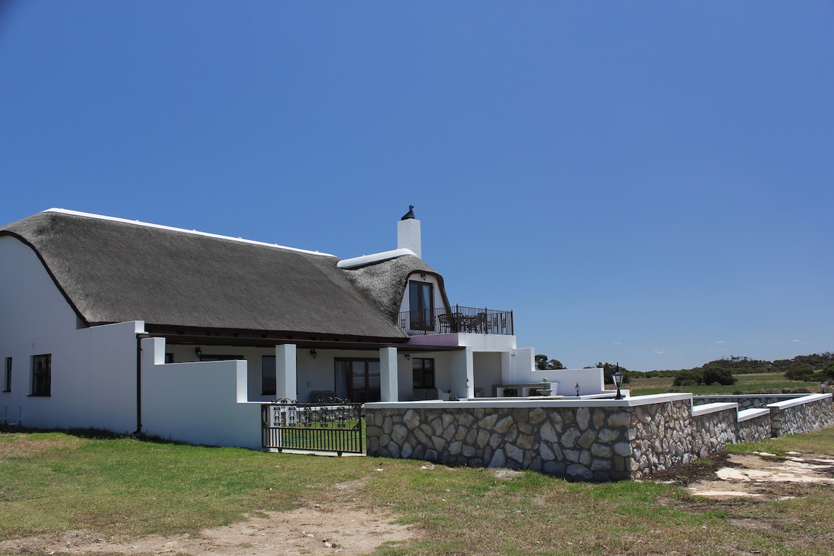 A restored thatch farmhouse is set against a clear blue sky, surrounded by a sprawling lawn. The stone wall features an entry gate, while large windows and a chimney create a charming facade. The outdoor area offers ample space for relaxation and enjoyment.