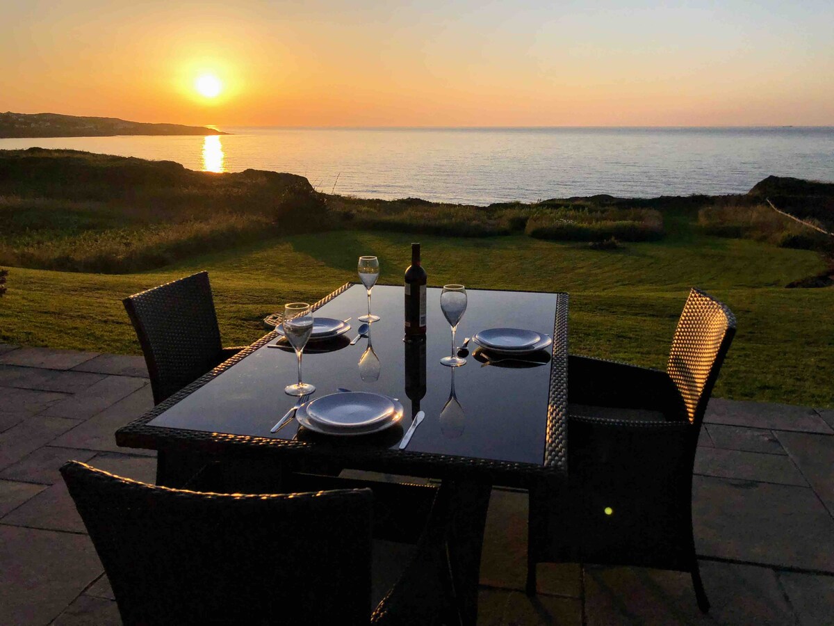 An outdoor dining table set with four chairs overlooks the sea at sunset. Plates and glasses are arranged for an evening meal, with the sun casting a warm glow on the tranquil water and lush green lawn beyond.