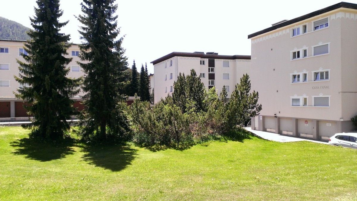 The image features a grassy area surrounded by trees and shrubs, providing a natural landscape. Two multi-story buildings can be seen in the background, characterized by their neutral-colored facades and simple architectural design, set against a clear blue sky.