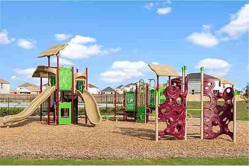 A colorful playground features various play structures, including slides and climbing walls. The ground is covered with mulch, enhancing safety. Surrounding the playground are green grass areas and a fence, with houses visible in the background under a clear blue sky.