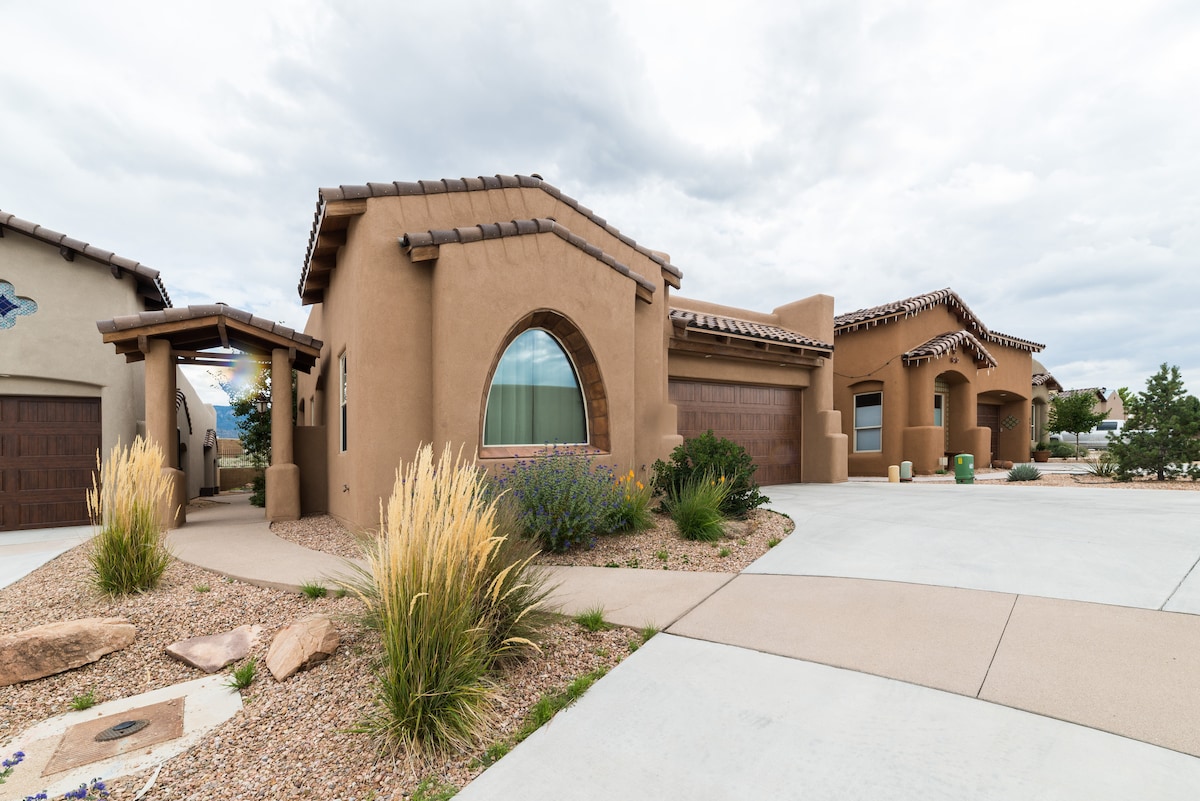 An elegant single-level home features a distinctive adobe-style exterior, characterized by arched windows and a tiled roof. Landscaping includes low-maintenance desert plants, with a decorative stone path leading to the entrance. An attached garage is visible on the right side of the image.