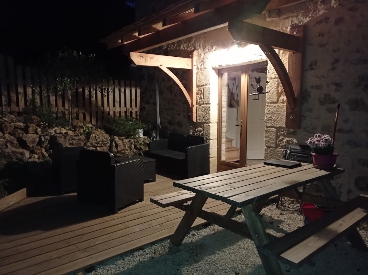 An outdoor seating area is presented, featuring a wooden picnic table and black wicker chairs arranged for comfort. Soft lighting from the entrance illuminates the stone facade of the house, while potted plants add a touch of greenery. A wooden fence borders the space.