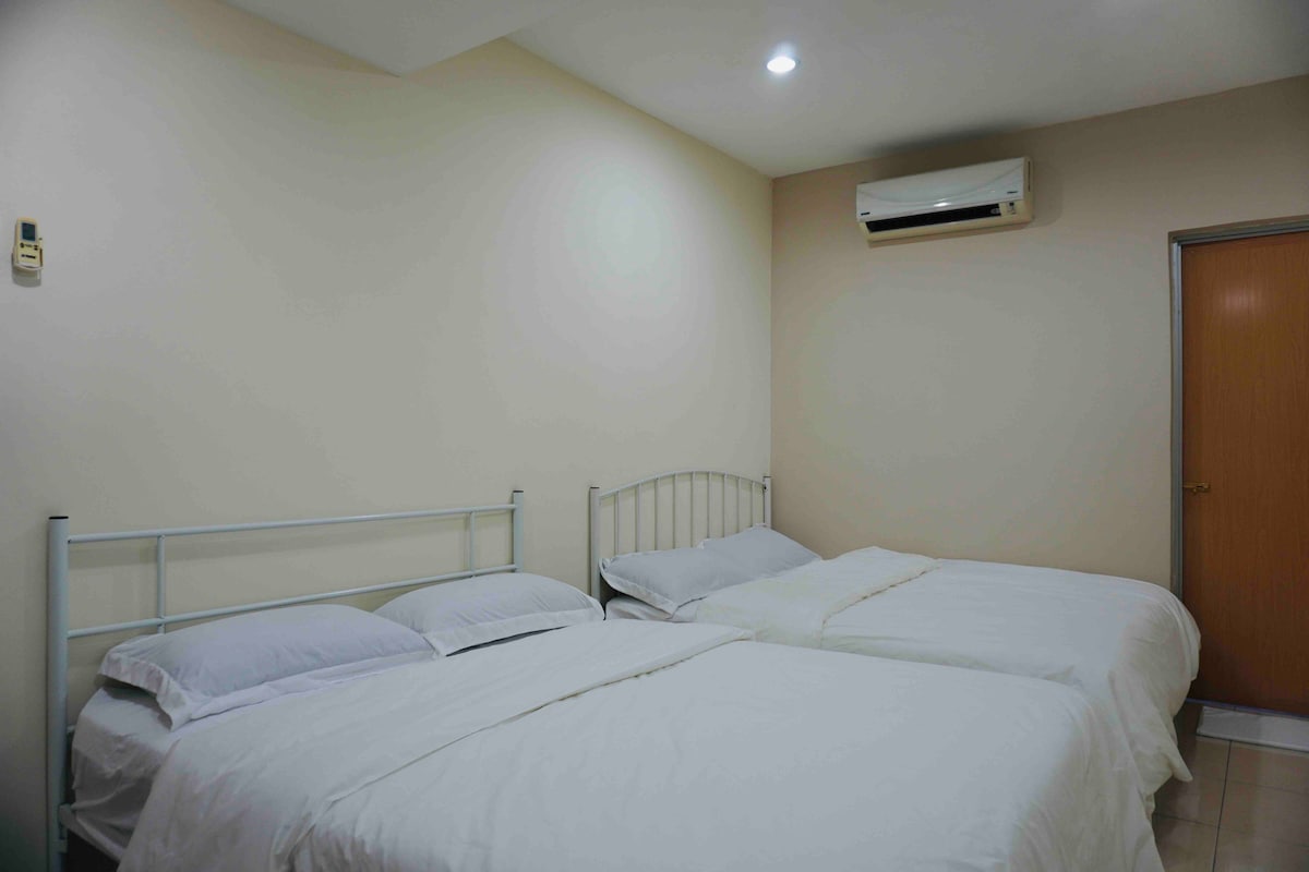 The sleeping area features two spacious beds, each with fresh white linens. Soft lighting from the ceiling illuminates the room, which has a neutral-colored wall. An air conditioning unit is mounted above, providing comfort. A wooden door leads to another area within the property.