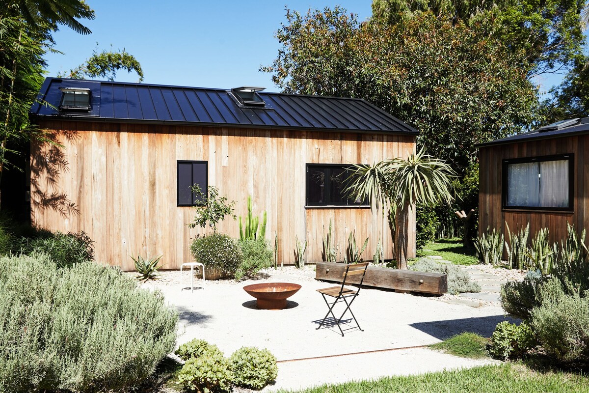 A wooden exterior structure with a black metal roof is surrounded by landscaped gardens featuring various plants. A circular fire pit sits on a gravel area, accompanied by a single black chair. Bright blue skies add depth to the serene outdoor setting.