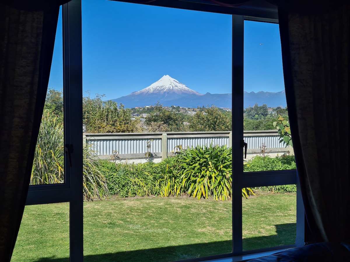 A clear view of Mount Taranaki is framed by large windows, showcasing a lush green lawn and garden in the foreground. Bright blue skies and snow-capped peaks create a tranquil backdrop, enhancing the natural beauty of the surroundings.