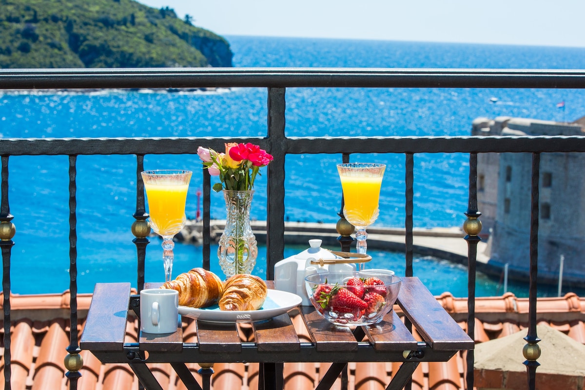 A balcony table is set with two glasses of orange juice and a plate featuring croissants and strawberries. A floral arrangement adds a touch of color, while the serene sea view provides a stunning backdrop of vibrant blue waters and the distant coastline.