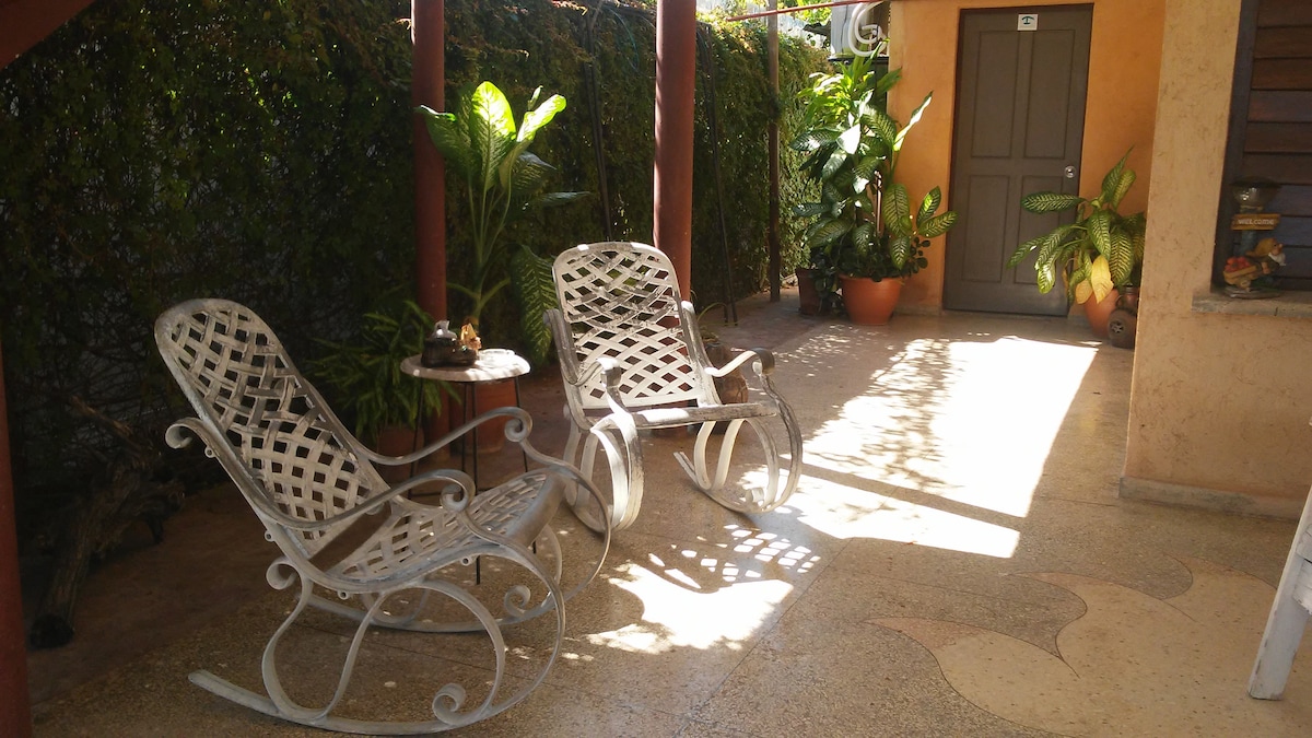 A shaded outdoor seating area features two white rocking chairs positioned beside a small round table. Lush green plants in terracotta pots line the space, and a door is visible in the background, providing an inviting atmosphere.