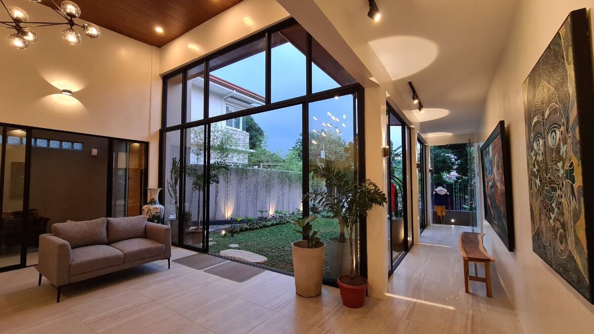 A spacious entryway is highlighted by large glass doors that invite natural light, connecting the indoor area to a lush garden. A comfortable sofa is positioned along the wall, accompanied by potted plants. Artistic wall decor adds visual interest to the space.
