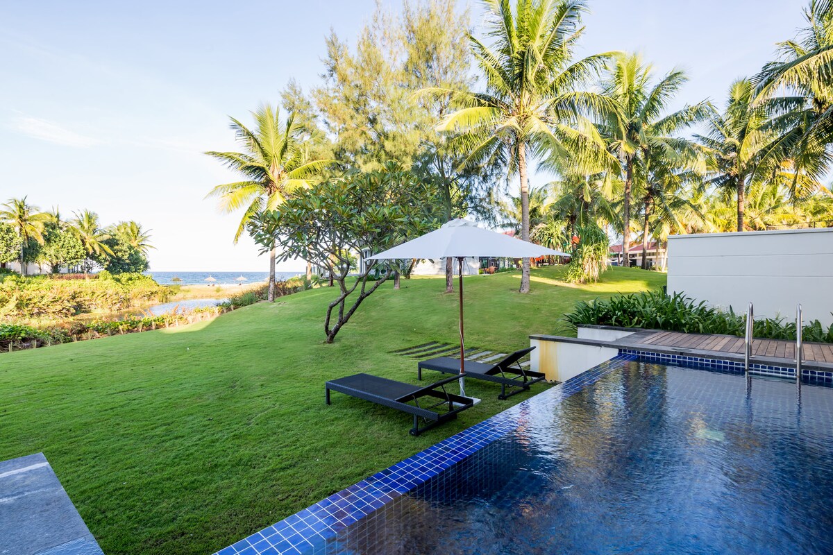 A refreshing private pool is set amidst a lush green lawn, with sun loungers positioned nearby for relaxation. Tall palm trees sway gently in the background, and a white umbrella provides shade. The sea is visible just beyond the landscaped garden.