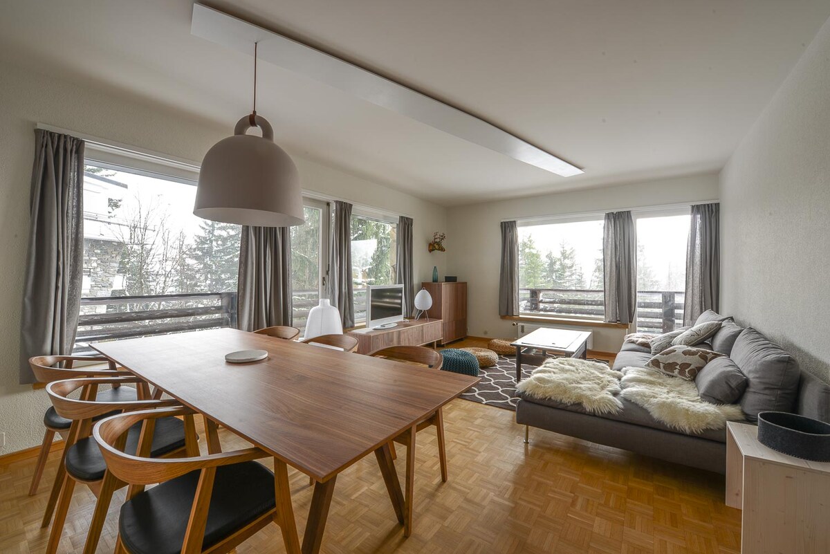 A spacious living area features a modern wooden dining table surrounded by stylish chairs. The cozy seating area includes a large sofa with decorative cushions, set against bright windows that provide natural light. Simple decor and natural tones enhance the inviting atmosphere.