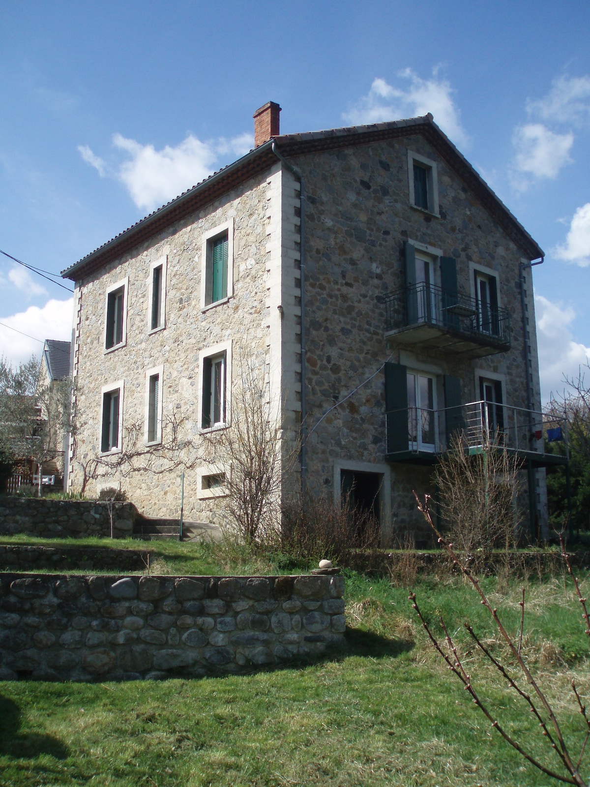 The exterior of a three-story stone house is captured under a bright blue sky, showcasing green shutters and a small balcony. Lush greenery surrounds the structure, with a terraced garden leading to the entrance. The rustic stone facade adds a charming character to the property.